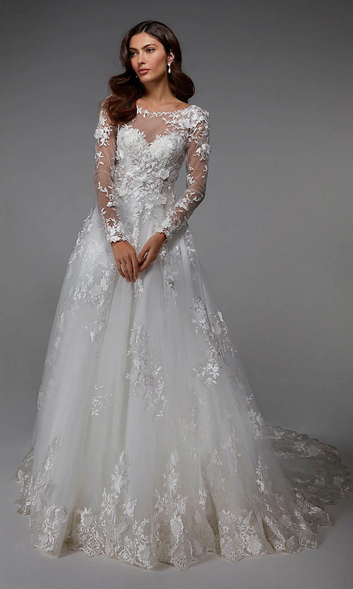 A woman stands against a plain gray background wearing the Alyce 7046 long ivory dress, featuring lace detailing, floral embroidery, sheer neckline, and long sleeves.